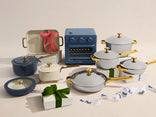 A collection of cookware, including pots, pans, a blue mini oven, a baking dish with a pink bow, and wrapped gift boxes with Our Place ribbon, arranged on a neutral background.