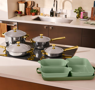 A set of stainless steel cookware with gold handles and lids, alongside a matching green ceramic dinnerware set, displayed on a modern kitchen countertop.