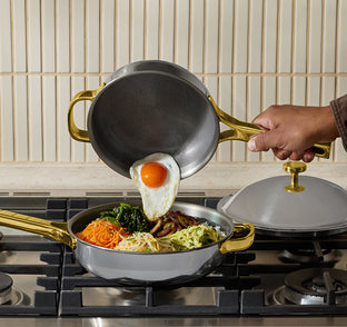 A hand pours a sunny-side-up egg from a small pan into a larger pan filled with mixed vegetables on a stovetop, with a pot lid resting nearby.