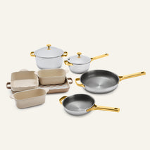 The Titanium Pro Cookware + Bakeware Set 2.0 includes two lidded pots, two frying pans, three baking dishes, and a checkered tray, all featuring metallic gold handles and knobs.