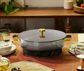 A gray lidded pot with gold handles sits on a dark trivet at the center of a set dining table with plates, glasses, and folded napkins.