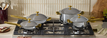 Four stainless steel pots and pans with gold handles and lids are arranged on a gas stove in a kitchen, surrounded by utensils, herbs, and cooking ingredients.
