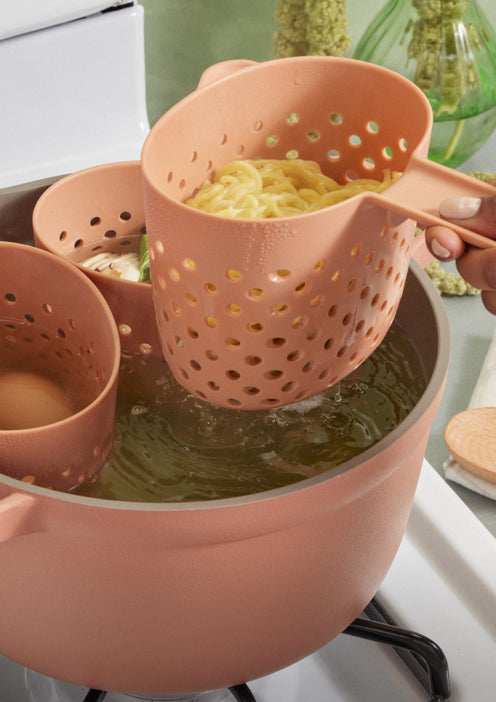 A hand uses a pink plastic strainer basket to lift cooked noodles from a pot of boiling water on a stove.
