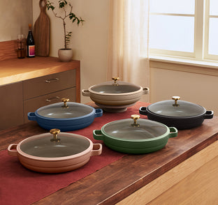 Five shallow pans with glass lids and gold handles in various colors are arranged on a kitchen island with a red runner.