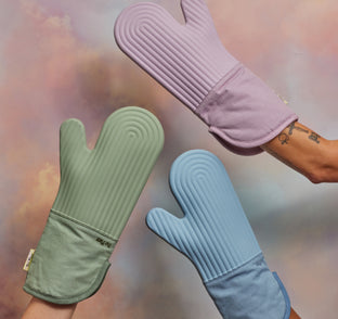 Three hands hold up silicone oven mitts in green, lavender, and blue against a soft, pastel-colored background.