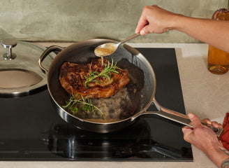 Person cooking steak in a pan on a stove with a bottle of wine in the background.