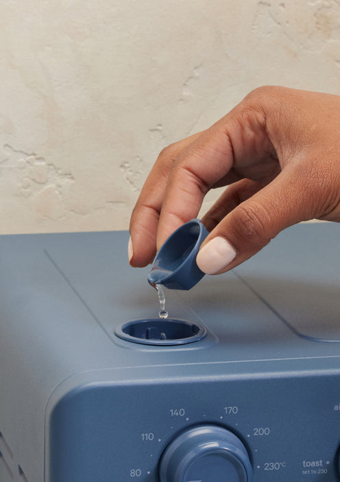 Hand pouring liquid into a blue appliance with a neutral background