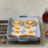 blue griddle pan on stove heating pancakes, eggs and bacon