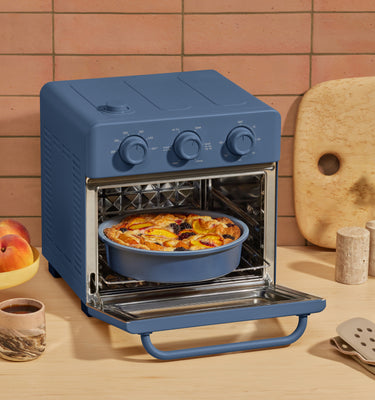 Blue toaster oven on a kitchen counter with food inside, surrounded by fruits, glasses, and a cutting board.