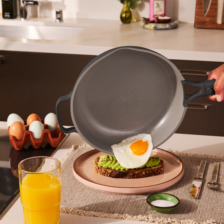 Person holding a frying pan with an egg on toast, next to a glass of orange juice in a kitchen setting.