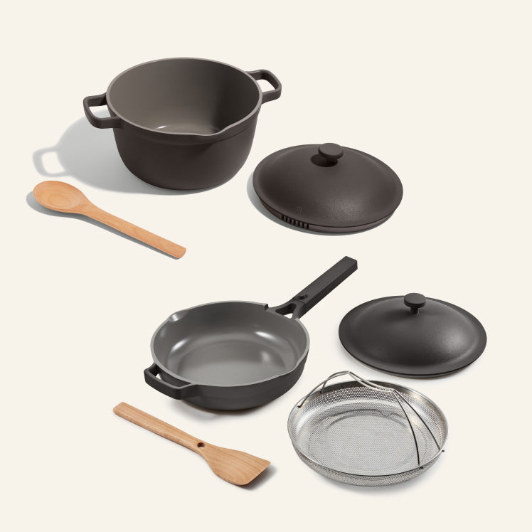 Set of black cookware including a wok, frying pans, lids, and a strainer on a light gray background.