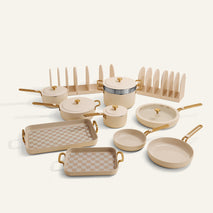 The Essentials Cookware + Bakeware Set in beige and gold includes pots, pans, bakeware, lids, and storage racks all arranged neatly on a light background.