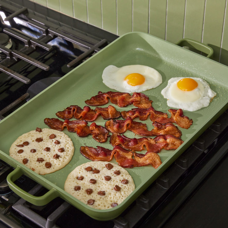 green griddle pan on stove cooking bacon, eggs, and pancakes