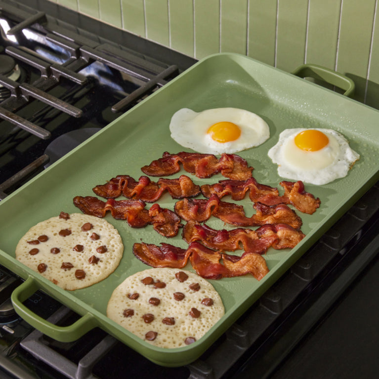 green griddle pan on stove cooking eggs, bacon and pancakes