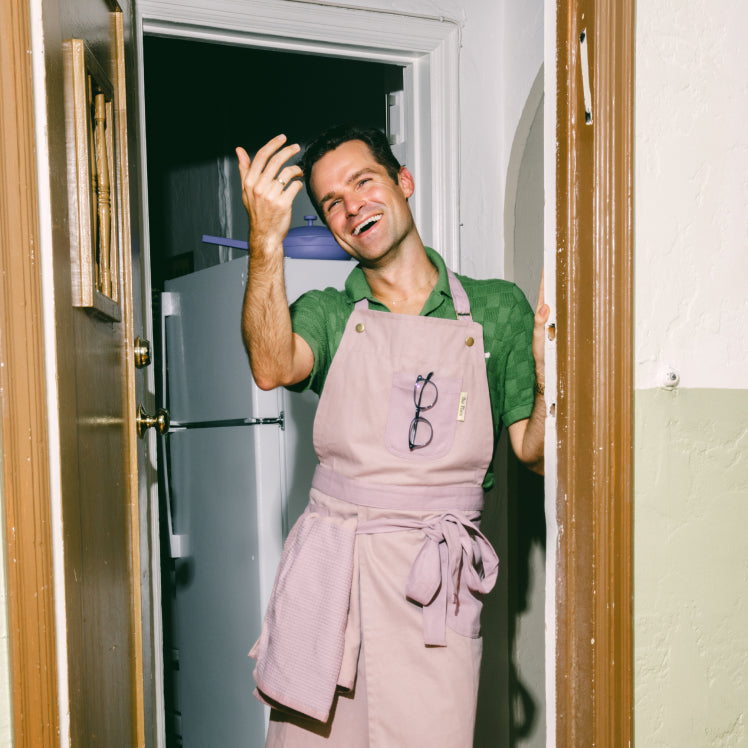 A chef wearing a purple Home Cook Apron raises a hand and smiles