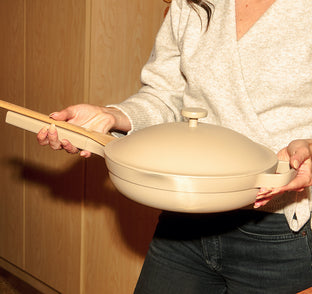 Person holding a beige ceramic pot with a wooden handle against a wooden background