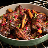 Braised lamb shanks with pomegranate seeds, rosemary sprigs, and garlic halves in a round pan, shown inside an oven.