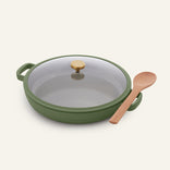 The Dual Handle Always Pan, featured in green with a nonstick surface and topped with a gold knob and glass lid, sits beside a light wooden spoon on a plain white background; crafted from non-toxic materials, this versatile pan is designed for healthier cooking.