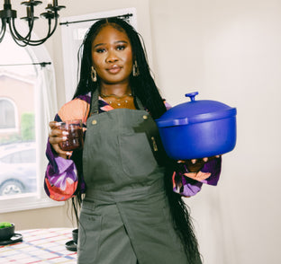Woman in an apron stands indoors holding a blue pot in one hand and a glass of red beverage in the other, with a table and chandelier in the background.