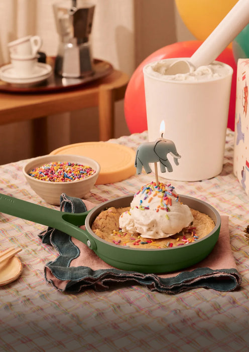 A skillet cookie topped with ice cream, sprinkles, and an elephant-shaped candle is on a table, with bowls of sprinkles and marshmallows, and colorful balloons in the background.