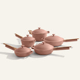 The Cookware Set+ includes five pink nonstick pieces with wooden handles: three saucepans with lids, a large pot with lid, and a frying pan, all arranged on a light surface.