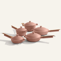 The Cookware Set+ includes five pink nonstick pieces with wooden handles: three saucepans with lids, a large pot with lid, and a frying pan, all arranged on a light surface.