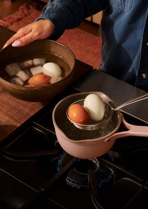 A person transfers boiled eggs from a saucepan on a stove into a bowl of ice water using a slotted spoon.