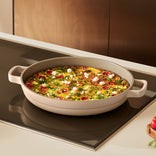 A round beige pan with handles holds a baked vegetable frittata with herbs and cheese, sitting on a stovetop in a modern kitchen.