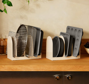 Two cream-colored organizers holding baking sheets, cooling racks, cake pans, and a muffin tin on a wooden countertop.
