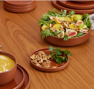 A wooden table with stacked brown dishes, a bowl of salad with figs and greens, a small plate of walnuts, and a bowl of yellow sauce.