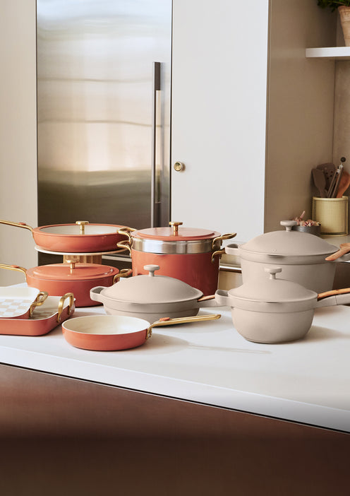 Assorted nonstick pots and pans in blush and beige colors with gold handles are arranged on a white kitchen counter.