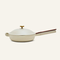 The Always Pan 2.0 is a cream-colored nonstick frying pan with a matching domed lid and long brown handle, part of a bestselling cookware system, shown against a plain light background.