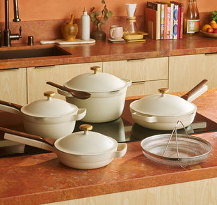 A set of four cream-colored pots and pans with lids and wooden handles on a kitchen counter, next to a wire strainer.