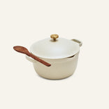 The Perfect Pot is an 8-in-1 cream-colored nonstick pot with a gold knob, lid, and a wooden spoon on the side handle, shown against a plain light background.
